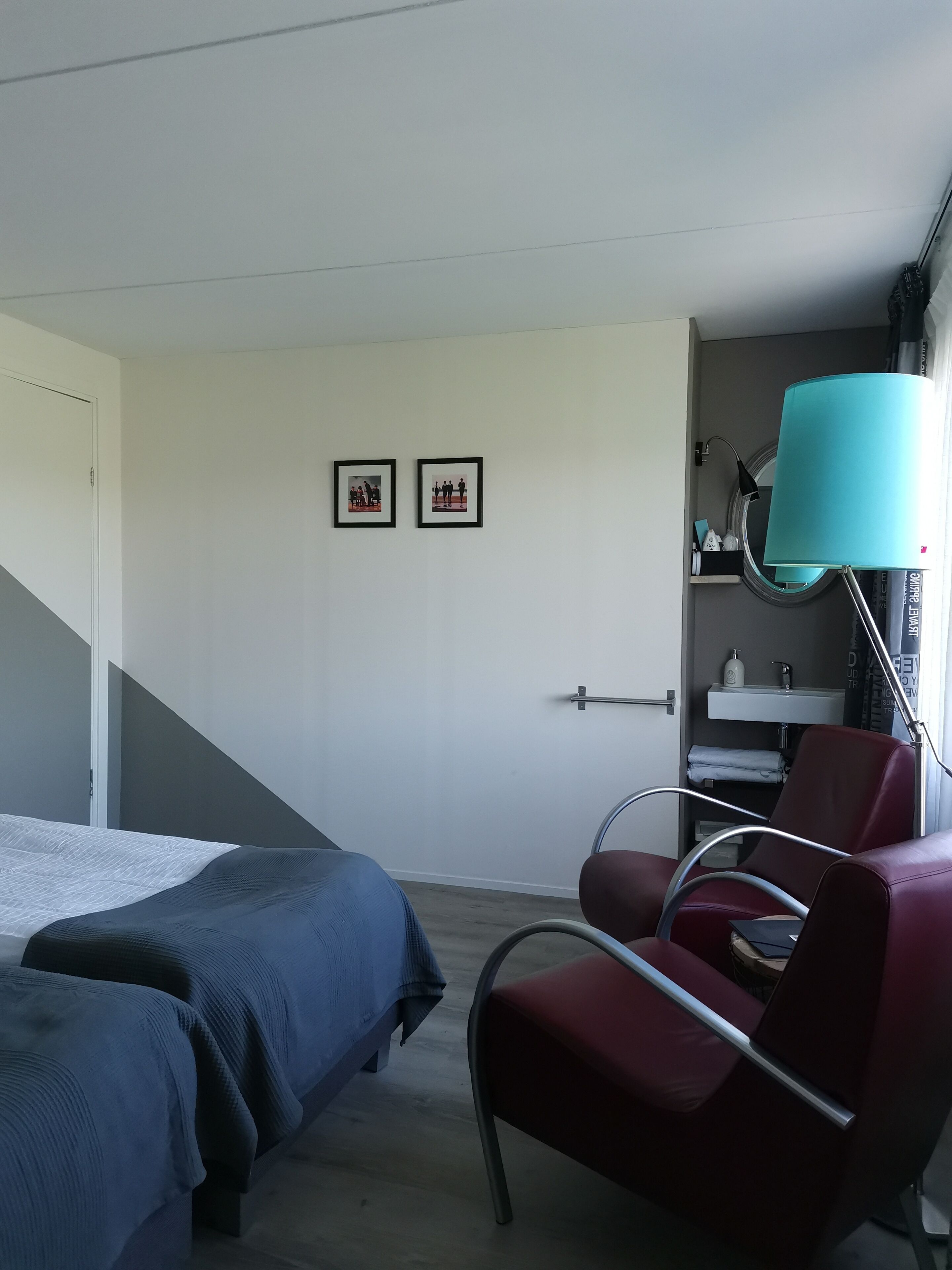 Una habitación de hotel con una cama, una silla roja, un escritorio con lámpara y dos cuadros en la pared.