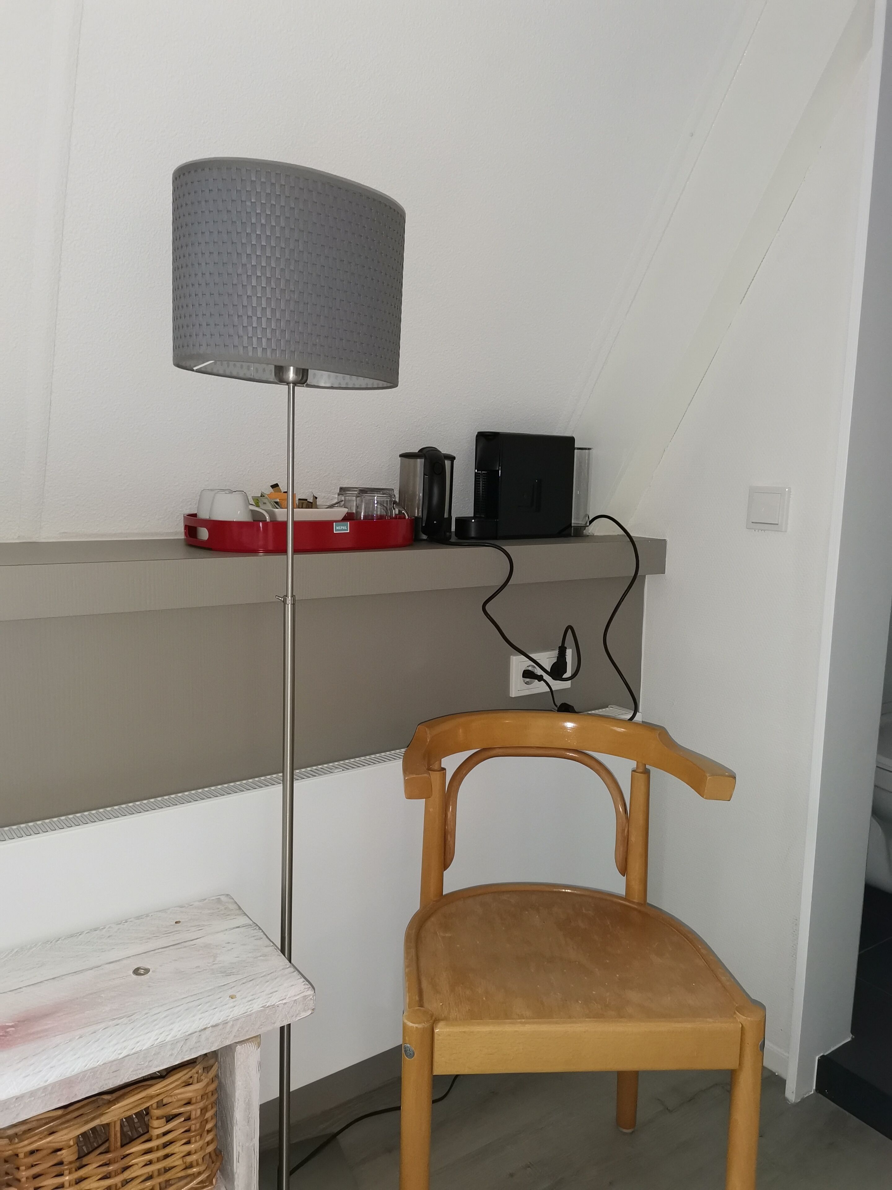 Una habitación pequeña con una silla de madera, una bandeja roja con utensilios de cocina, una cafetera negra y una lámpara.