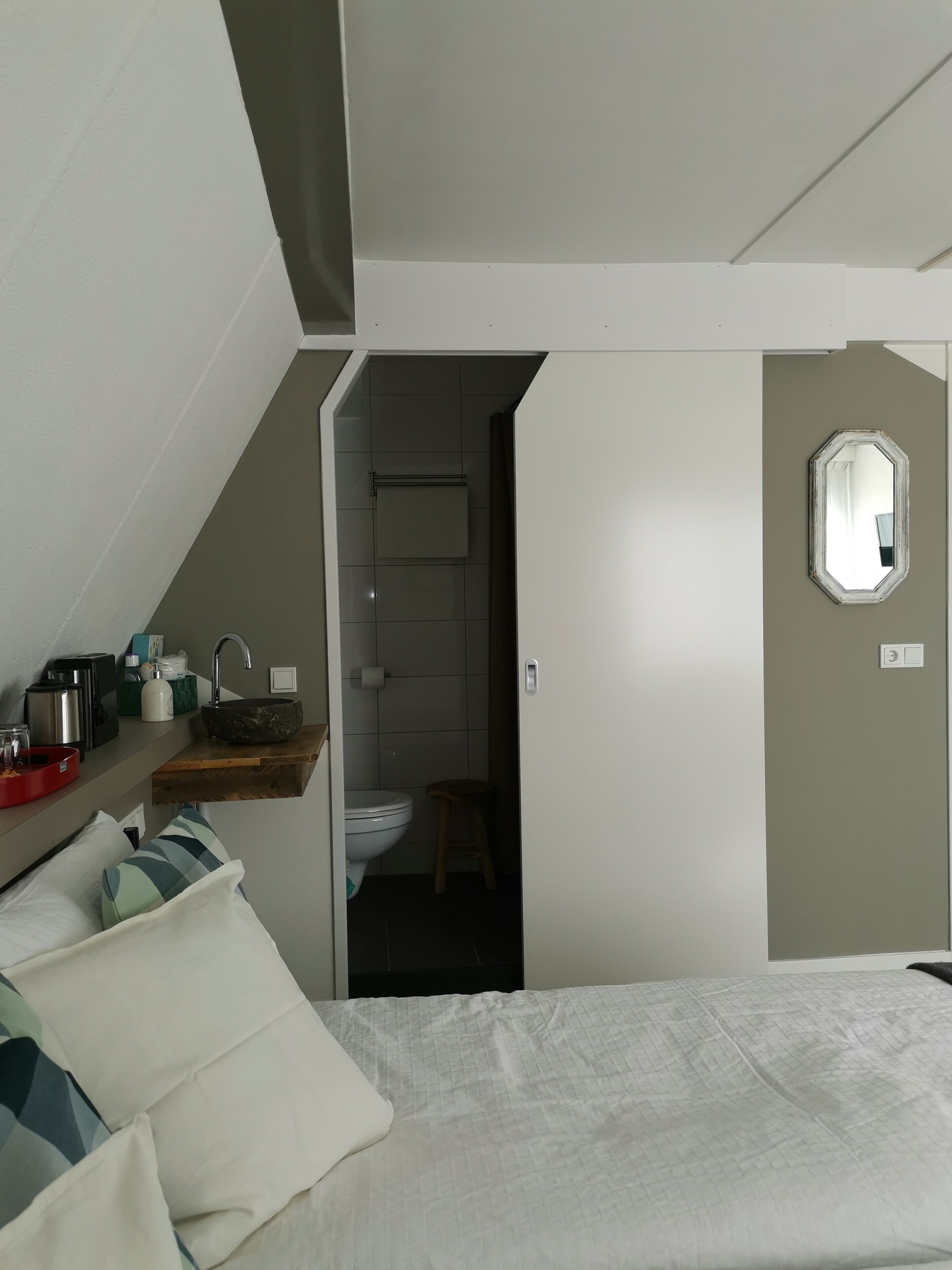 Een slaapkamer met een bed, een nachtkastje met een rode dienblad, een houten kruk, en een badkamer zichtbaar door een geopende deur.