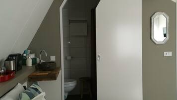 Een slaapkamer met een bed, een nachtkastje met een rode dienblad, een houten kruk, en een badkamer zichtbaar door een geopende deur.