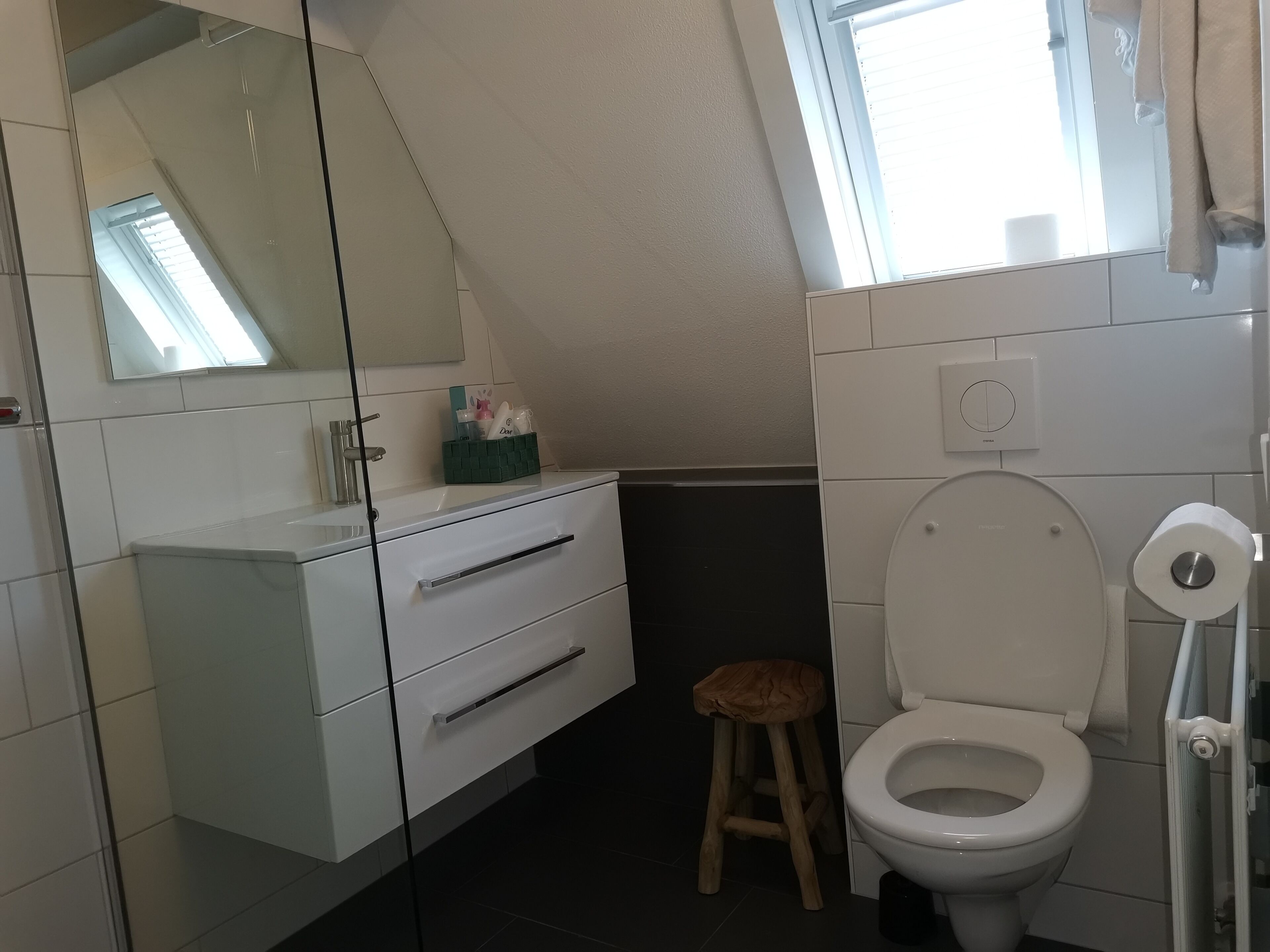 Ein modernes Badezimmer mit einem weißen Waschtisch, einem hölzernen Hocker, einer Toilette und einem Fenster mit Tageslicht.