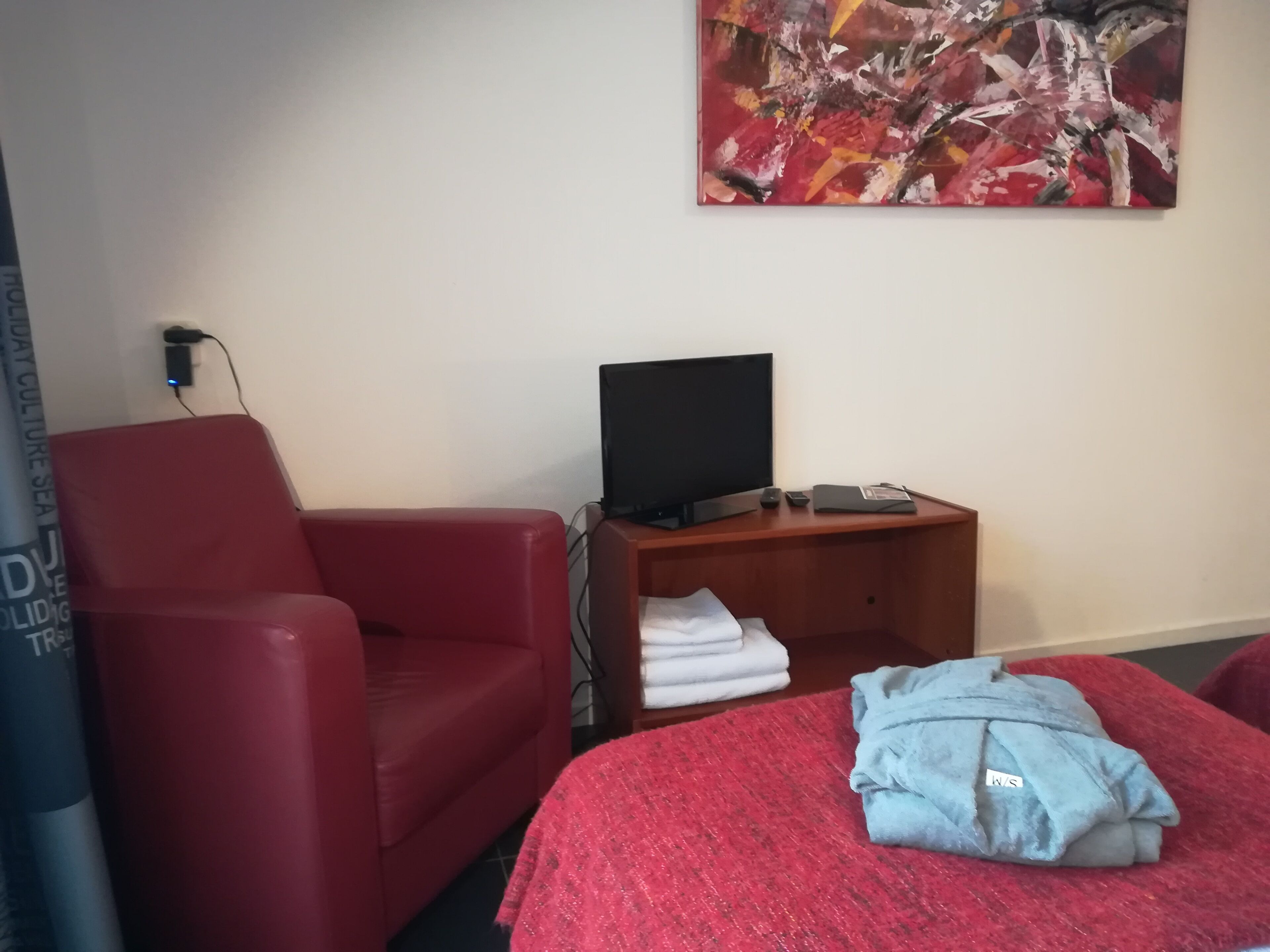 Een hotelkamer met een rode fauteuil, een televisie op een houten standaard en een bed met een rode sprei en een blauw kledingstuk.