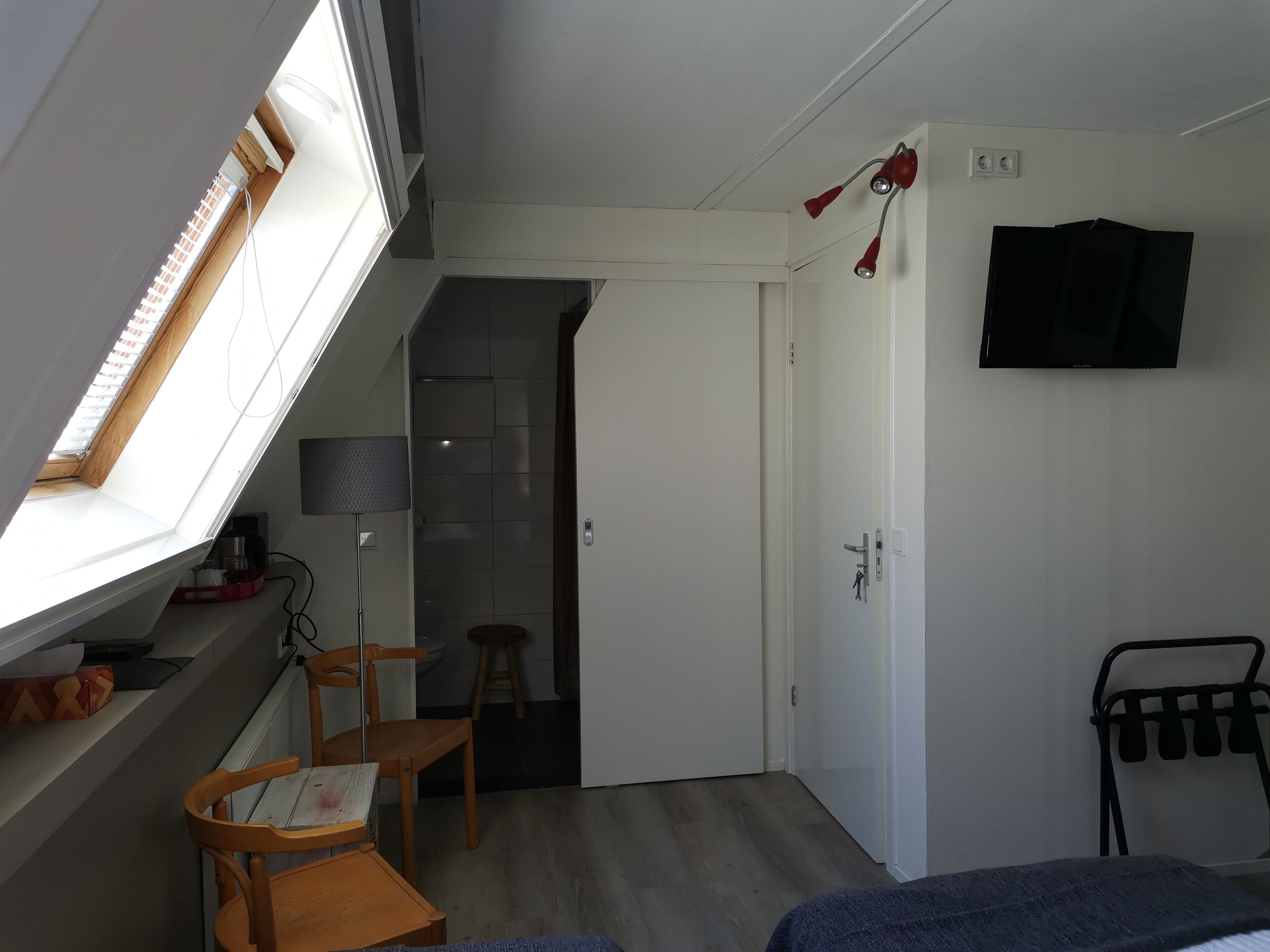 Une pièce de taille modeste, dotée d’une lucarne, d’un lit, d’un bureau avec une chaise, d’une télévision et d’une porte donnant sur une autre pièce.