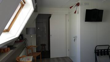 Une pièce de taille modeste, dotée d’une lucarne, d’un lit, d’un bureau avec une chaise, d’une télévision et d’une porte donnant sur une autre pièce.