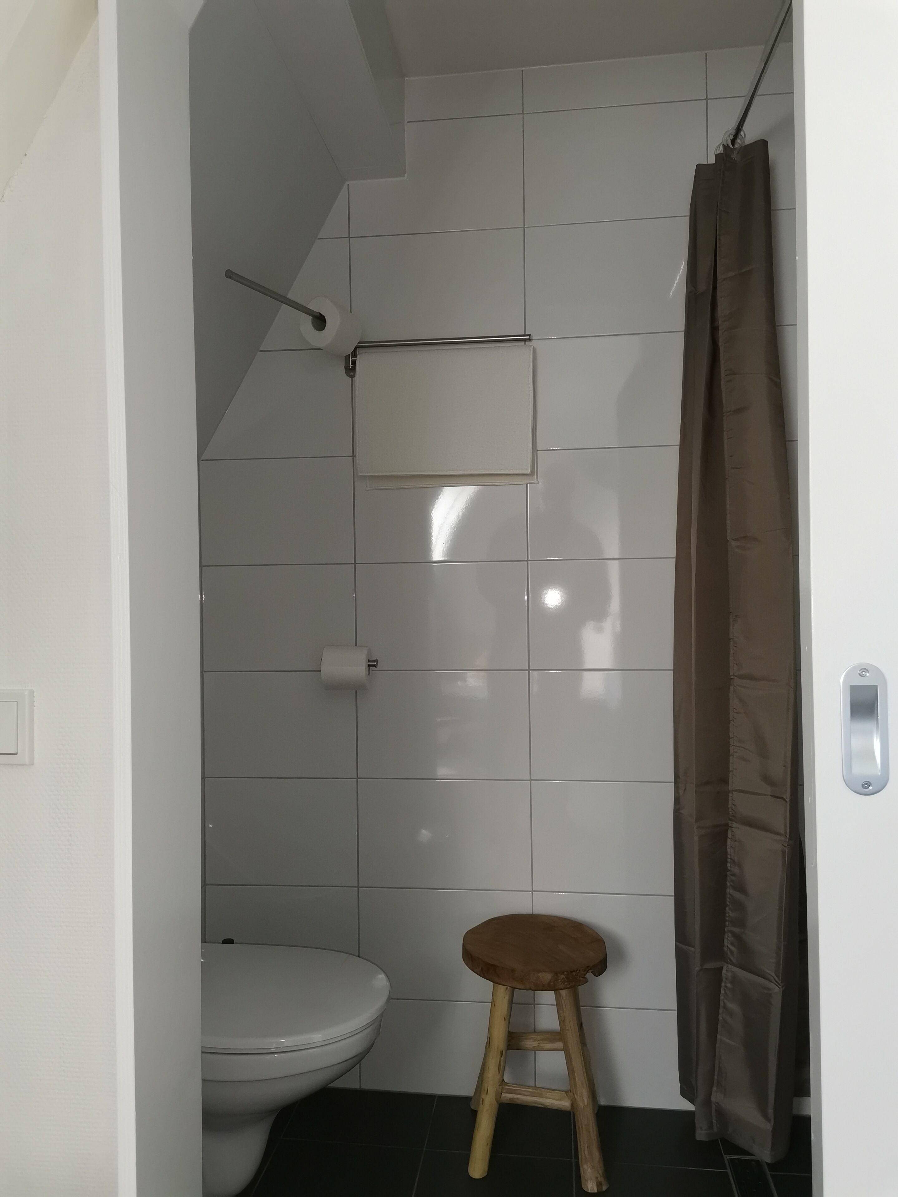 A small, tiled bathroom with a toilet, a wooden stool, and a shower curtain.