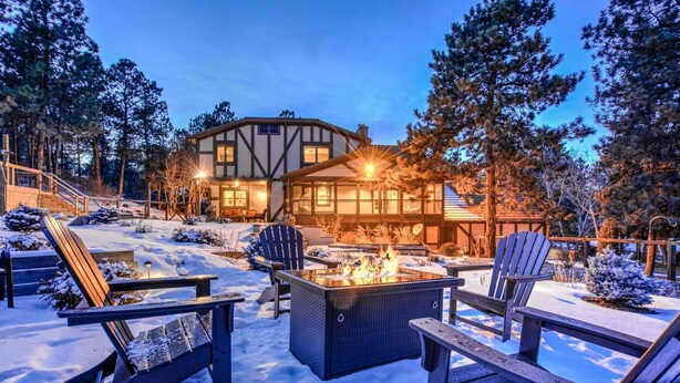 Un rincón exterior acogedor con sillones Adirondack y una chimenea, rodeado de nieve y una casa grande con una escalera visible.