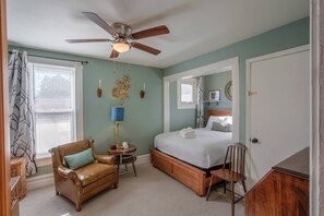Un dormitorio con cama, una silla, una lámpara, un ventilador de techo y una ventana con cortinas.