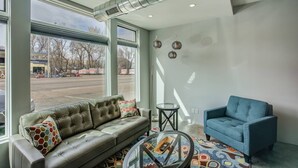Una sala de estar moderna con un sofá de cuero, un sillón azul y una mesa de centro de vidrio. Amplias ventanas ofrecen vista a la calle.