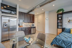 Un apartamento moderno con cocina, comedor y habitación. Incluye electrodomésticos de acero inoxidable, una mesa de comedor de vidrio y una cama con cubrecama azul.