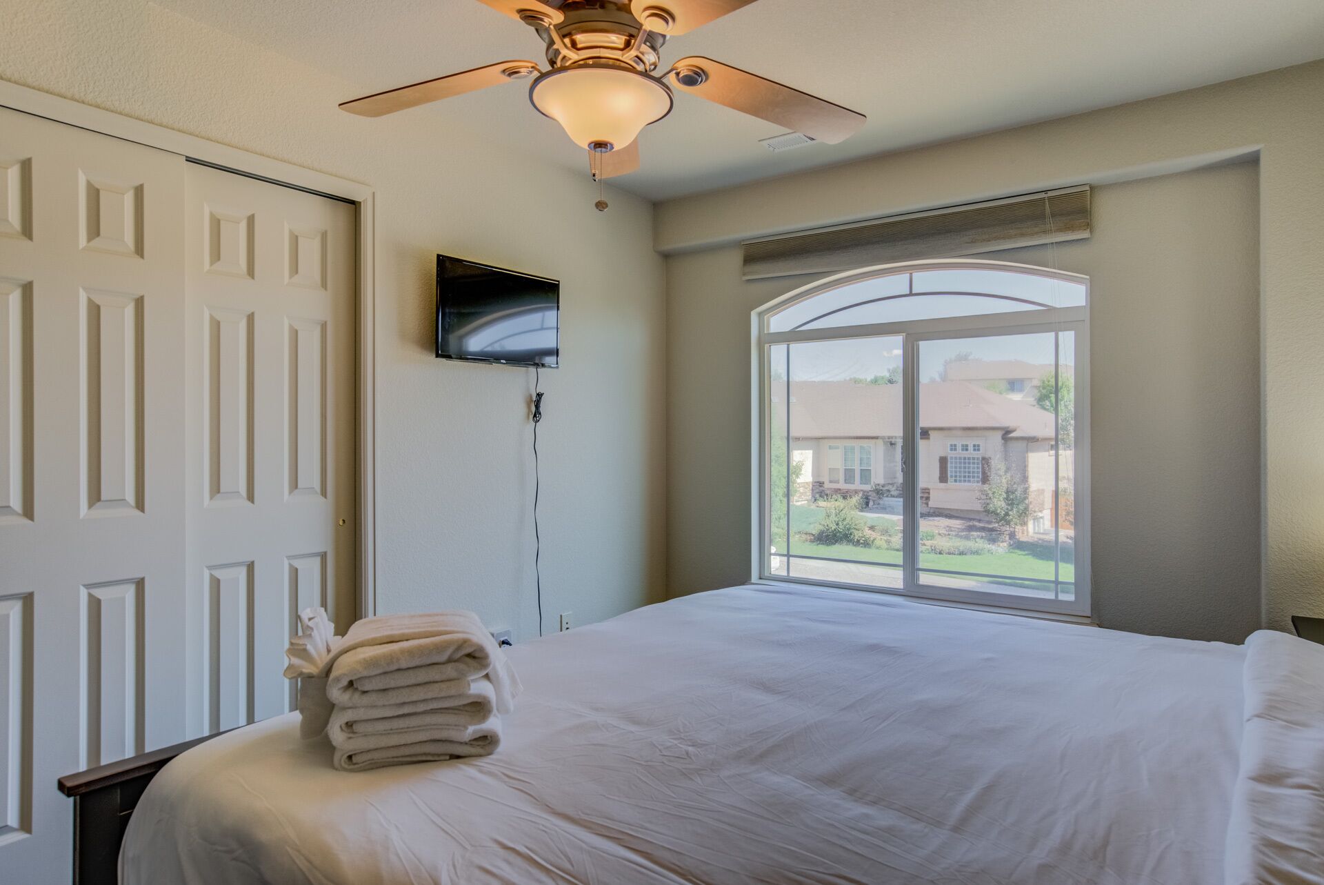 A bedroom with a bed, a ceiling fan, a TV mounted on the wall, and a window with a view of a house.