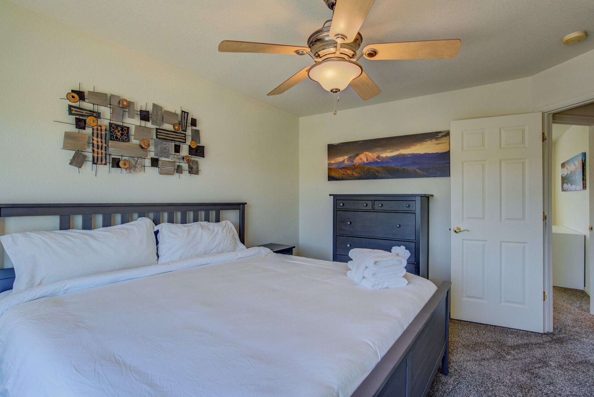 A neatly made bed with white linens, a ceiling fan, a wall art piece, a dark dresser, and a door leading to another room.