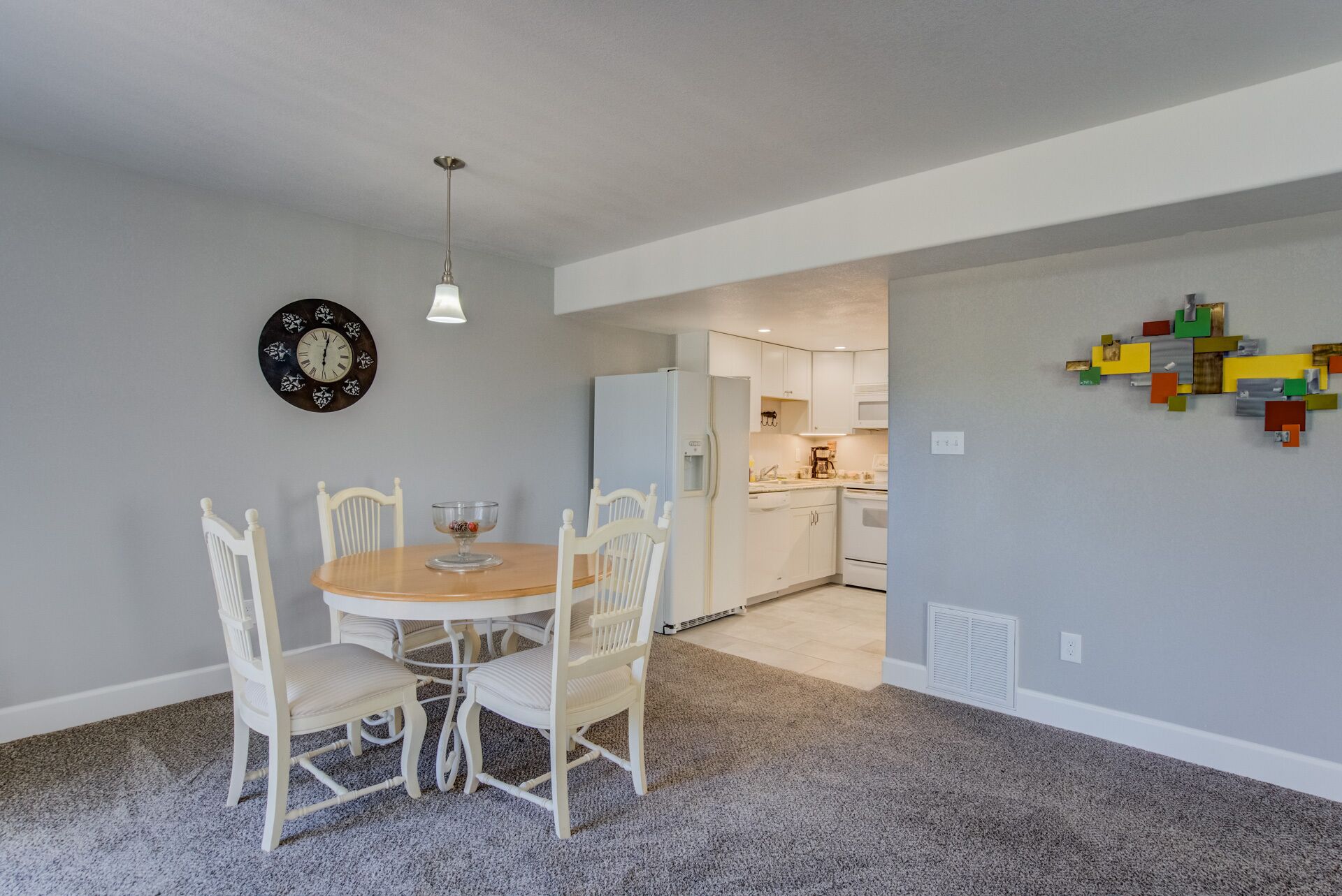 A dining area with a round table and four chairs, a wall clock, and a kitchen in the background.