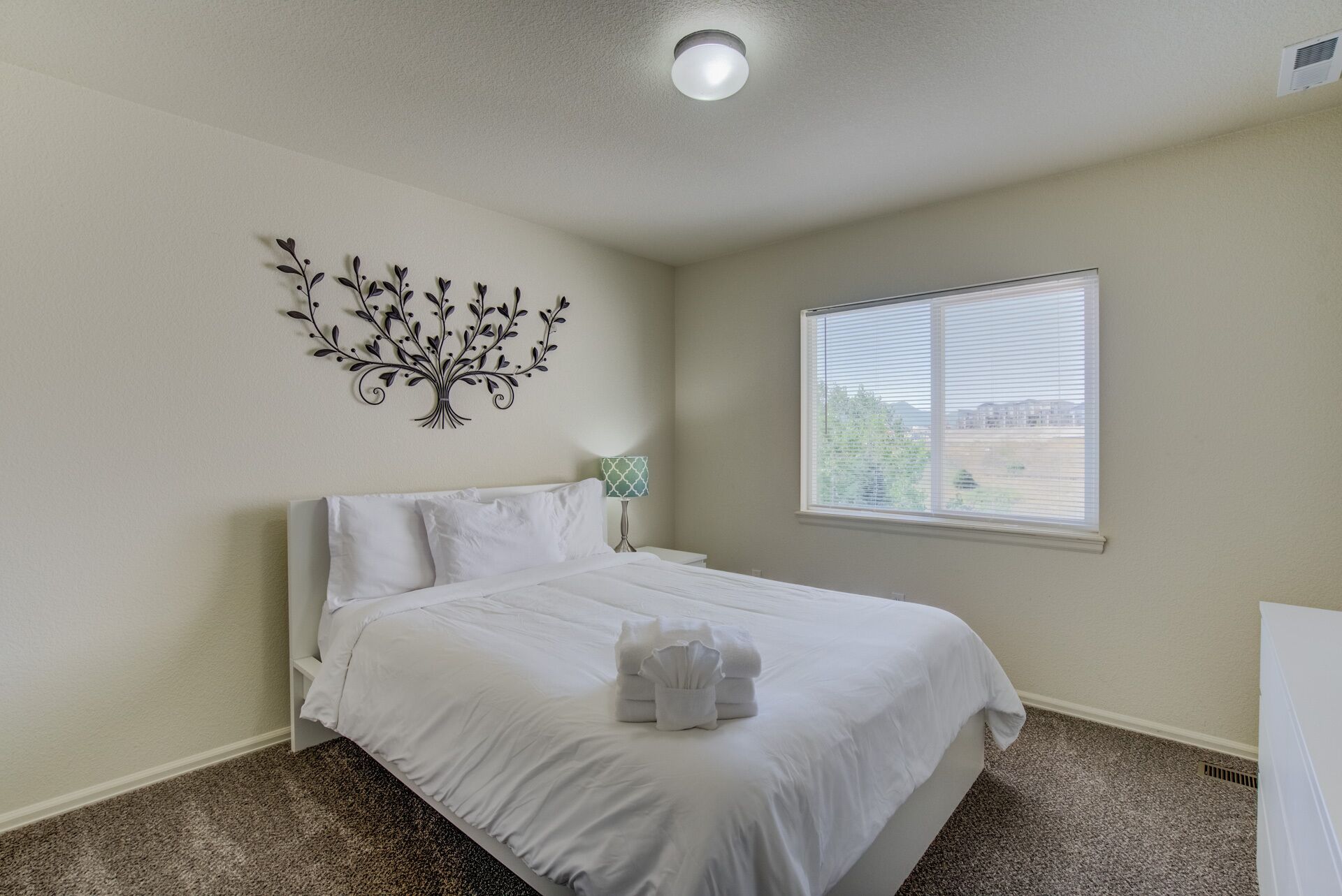 A bedroom with a bed, a wall-mounted decorative tree, a bedside lamp, and a window with a view.