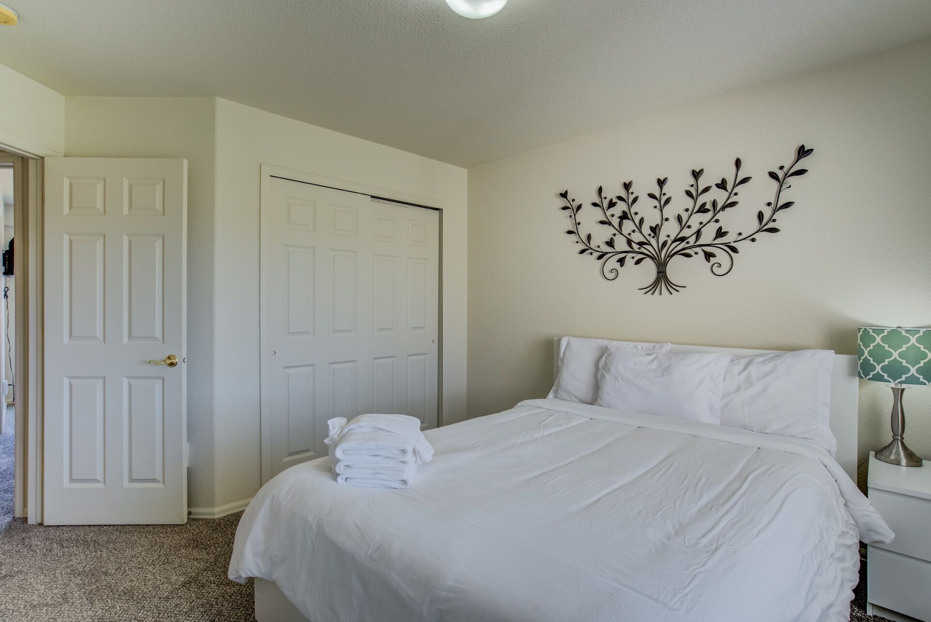 A neatly made bed with white linens, a wall-mounted decorative tree, a closed white door, and a bedside table with a lamp.