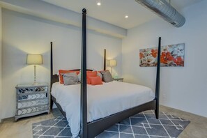 A four-poster bed with a white bedspread and multiple pillows, a nightstand with a lamp, a patterned rug, and a floral painting on the wall. - 3BR Modern Marveldowntown Customprivate Balcony (Colorado Springs)