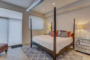 A four-poster bed with a white bedspread and multiple pillows, a bedside table with a lamp, and a patterned rug on the floor. - 3BR Modern Marveldowntown Customprivate Balcony (Colorado Springs)
