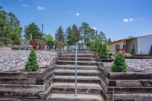 A set of wooden steps leading up to a metal pole, surrounded by small trees and a gravel area with Adirondack chairs. - Garden-level Perfect Hot tub Fire Pit & More! (Colorado Springs)