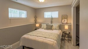 A bedroom with a bed, two bedside tables, a nightstand, a ceiling light, two windows with blinds, and a door leading to another room. - 2BR Garden-level Great Outdoors Dog Friendly (Colorado Springs)