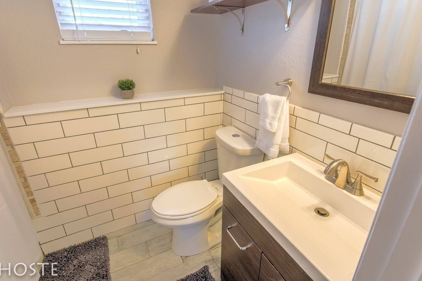 A bathroom with a toilet, sink, and mirror. There is a window with blinds and a small plant on the wall.