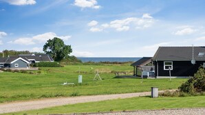 A rural landscape with a gravel path, a grassy field, and a body of water in the distance. There are two buildings with dark roofs and a large tree. - Beautiful holiday home on Nordals, Vinkelbæk 40 (Nordborg)
