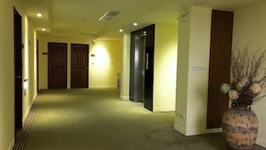 Un couloir d’hôtel avec des revêtements de sol en moquette, des portes en bois et un grand vase contenant des branches séchées.