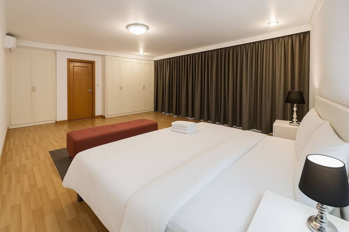 Una habitación de hotel moderna con una cama grande, un banco rojo, una mesita de noche con lámpara y un armario.