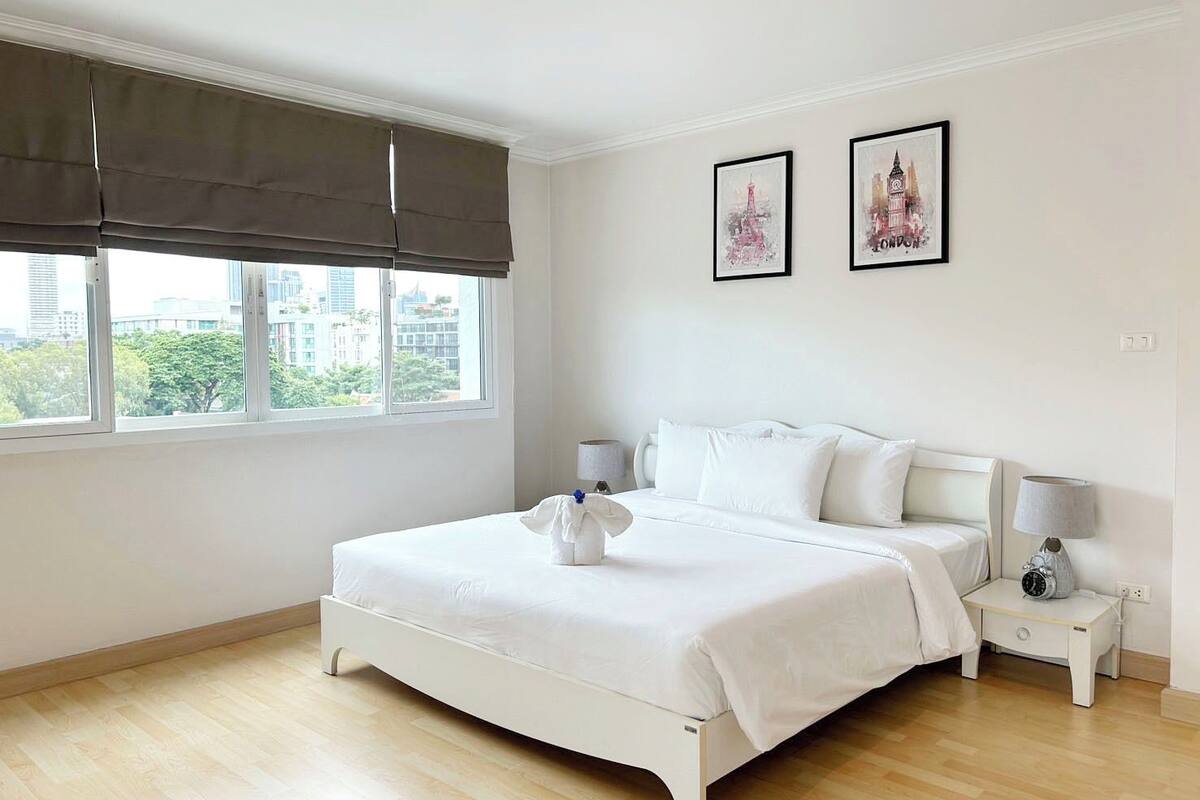 Un dormitorio con una cama, dos mesitas de noche, un ventanal con vistas a edificios y dos cuadros enmarcados en la pared.
