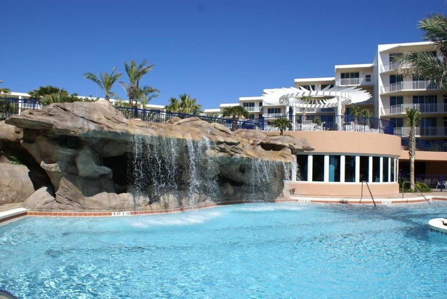 A pool with a waterfall feature, surrounded by a multi-story hotel with balconies and palm trees.