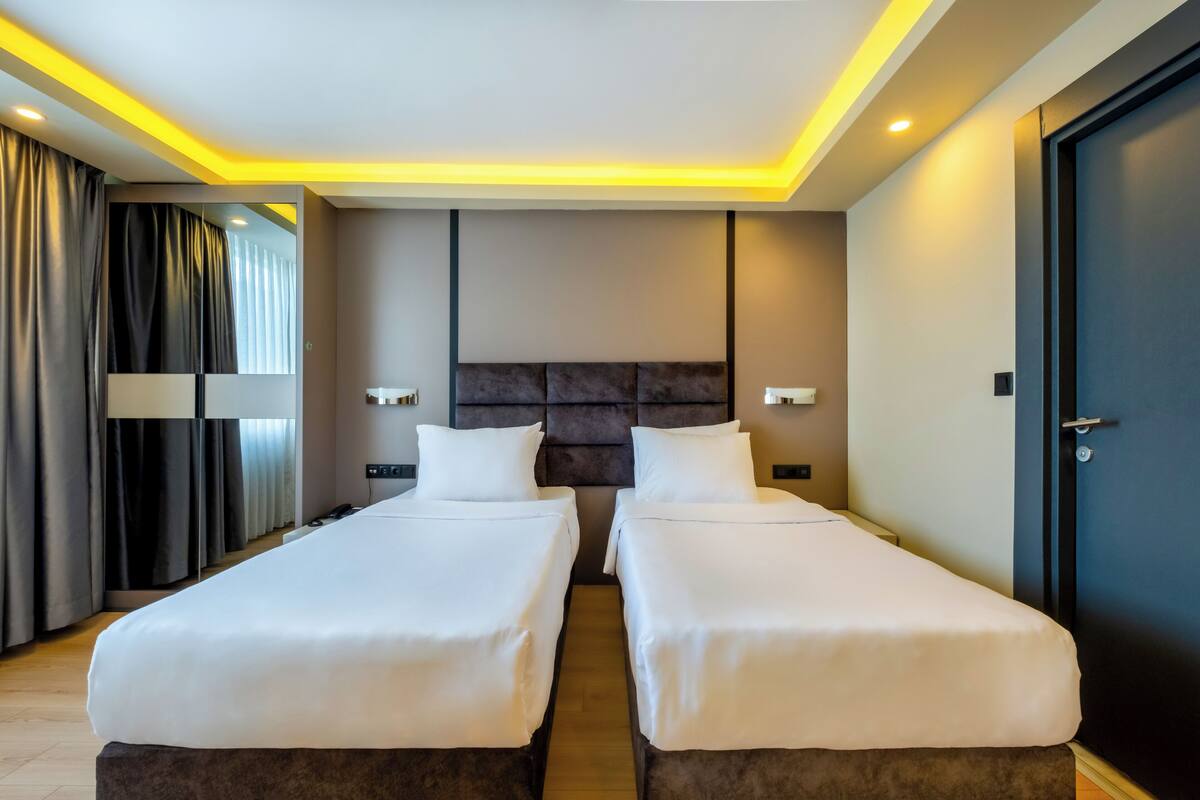 A modern hotel room with two single beds, a dark headboard, and a large window with curtains.
