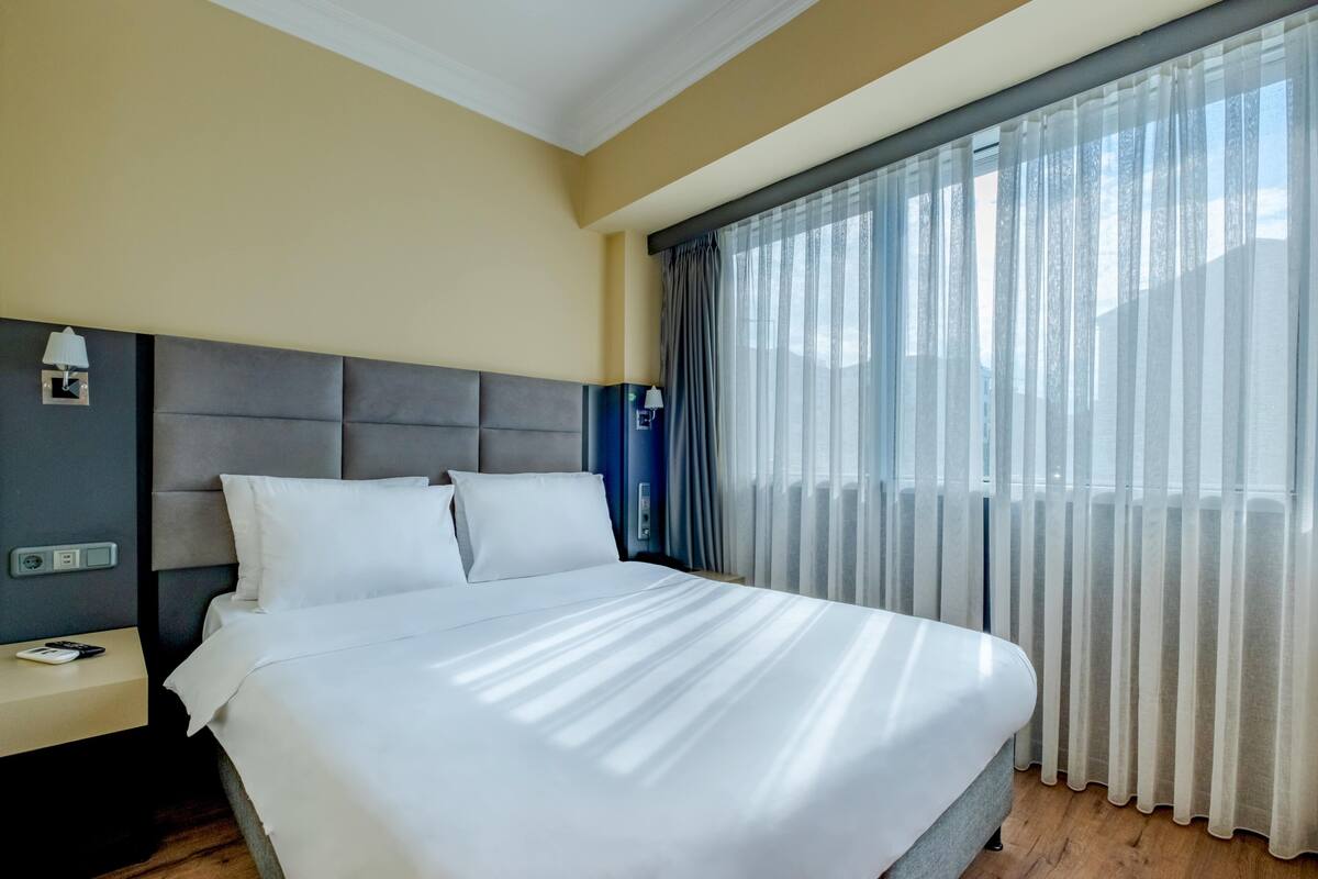 A hotel room with a large bed, a nightstand, a lamp, and a window with sheer curtains.