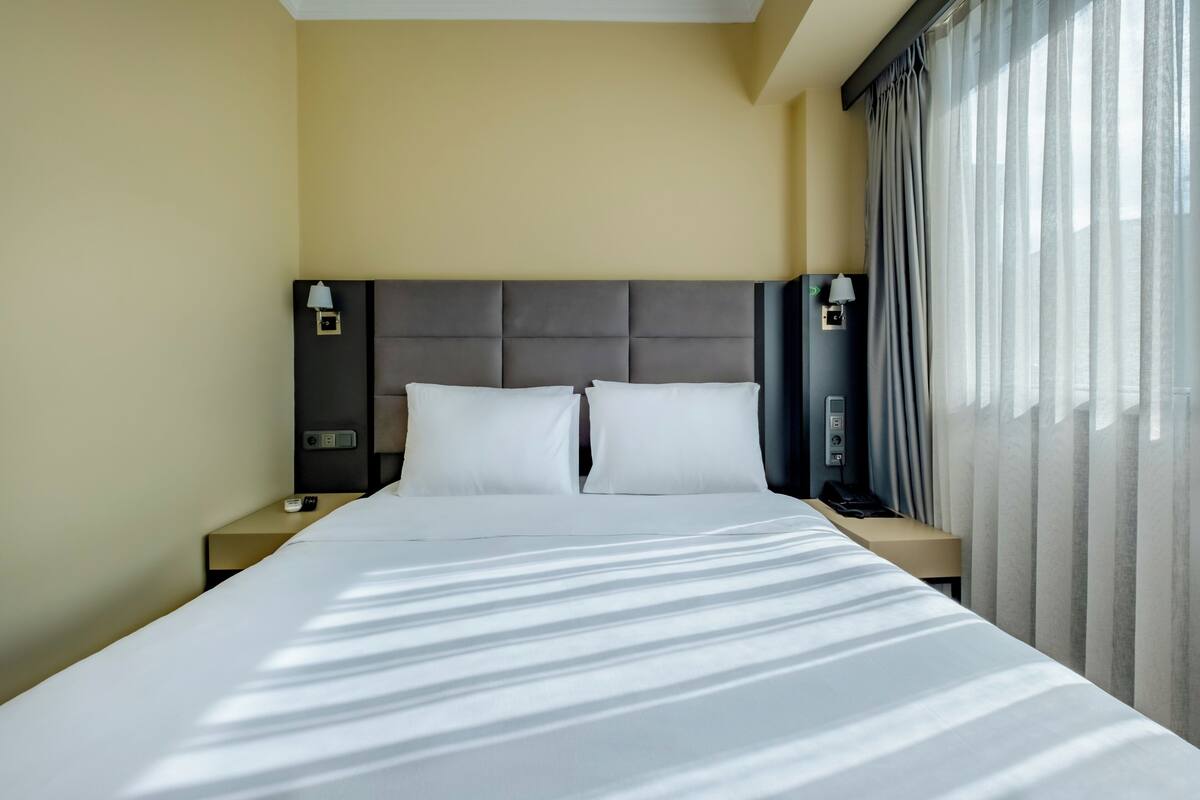 A hotel room with a large bed, two bedside tables, a headboard, and a window with curtains.