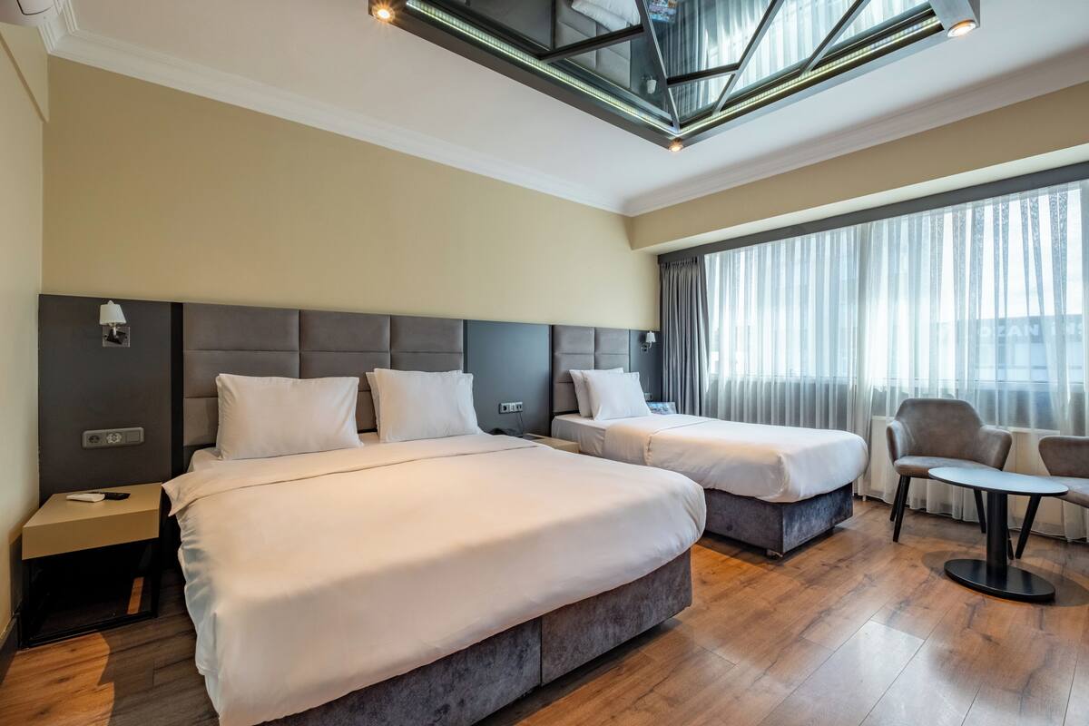 A hotel room with two beds, a small table, a chair, and a large window with curtains.