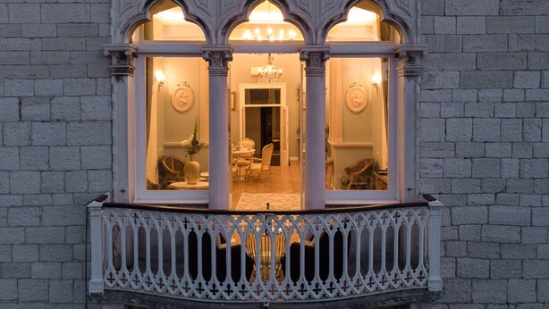 Ein Erker mit drei Rundbögen, ein Balkon mit aufwendiger Geländerkonstruktion und Blick in einen behaglich beleuchteten Innenraum mit Möbeln und einer Kronleuchter.
