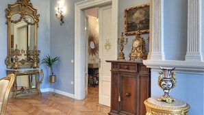 Ein Zimmer mit Fischgrätparkett, einem großen, verzierten Spiegel, einer Holzkabine mit Goldverzierungen und einem kleinen, runden Tisch mit einer Vase.