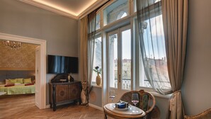 Ein Zimmer mit Fischgrätparkett, einer Couch mit grüner Auflage, einem Holzschrank, einem Fernseher und einem Fenster mit Vorhängen.