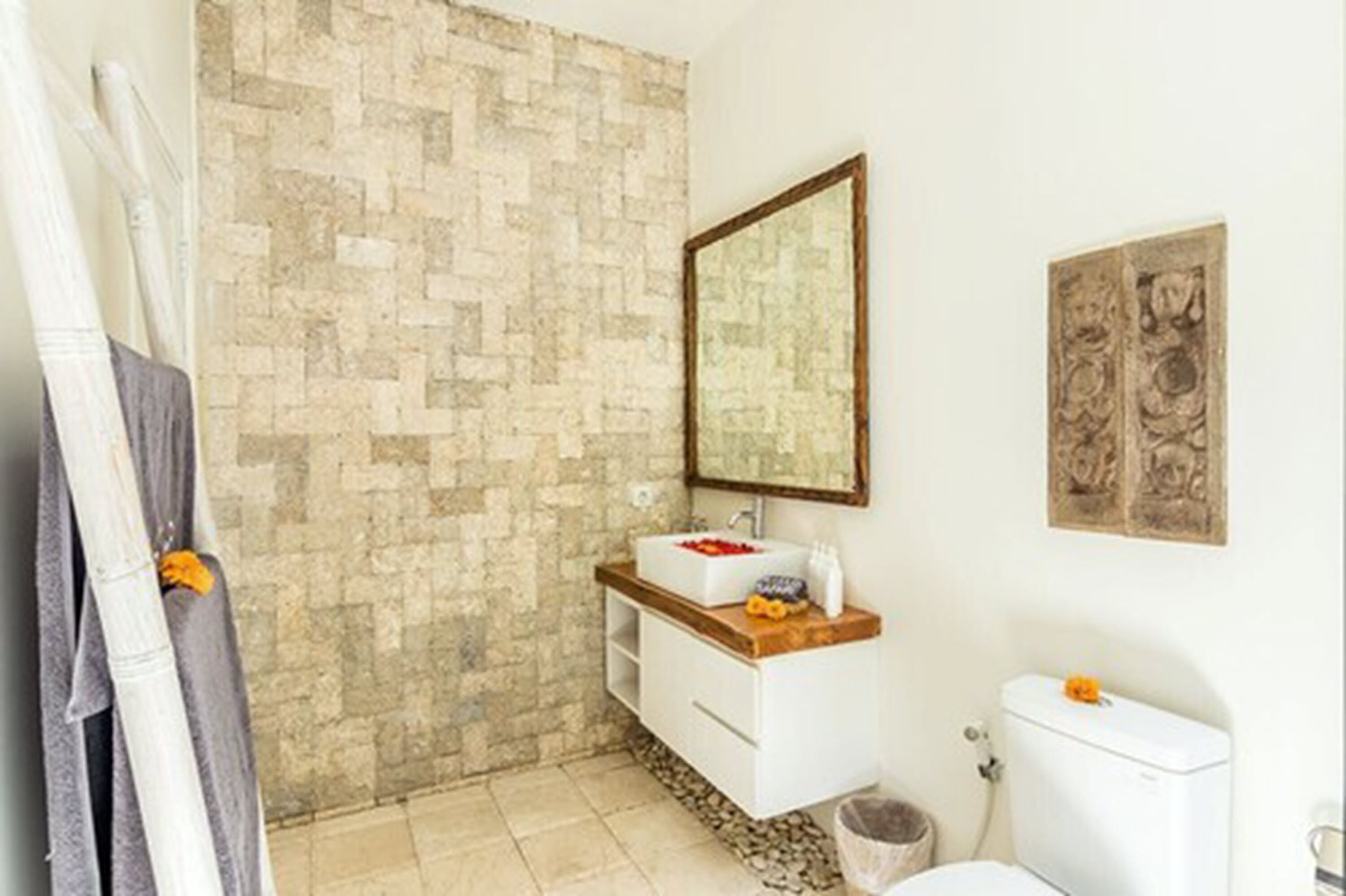 A bathroom with a stone wall, a white sink with a wooden countertop, a mirror above the sink, and a toilet.