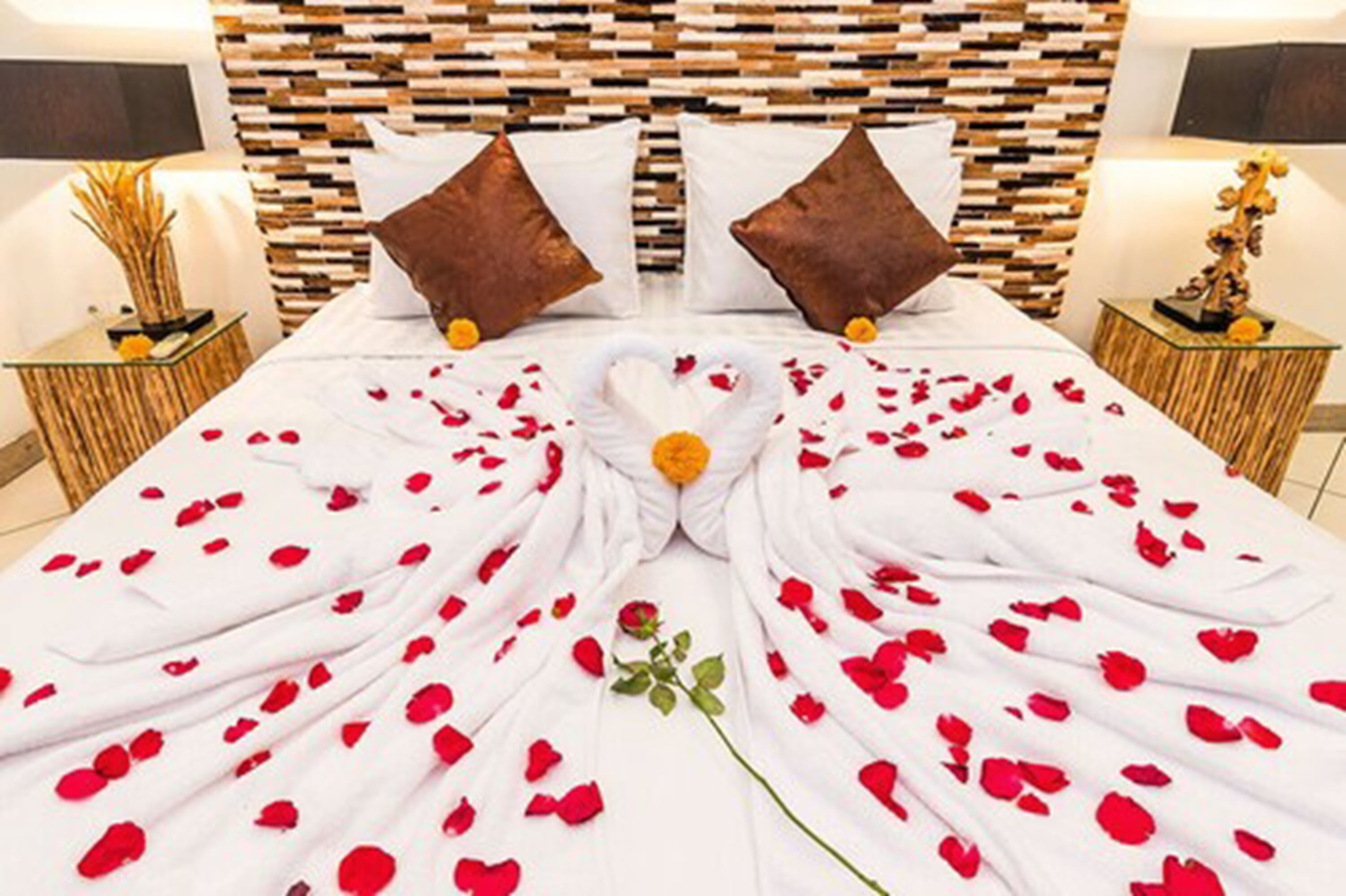 Una cama bien tendida con pétalos de rosa y una sola rosa, dos almohadas marrones y un cabecero de madera.