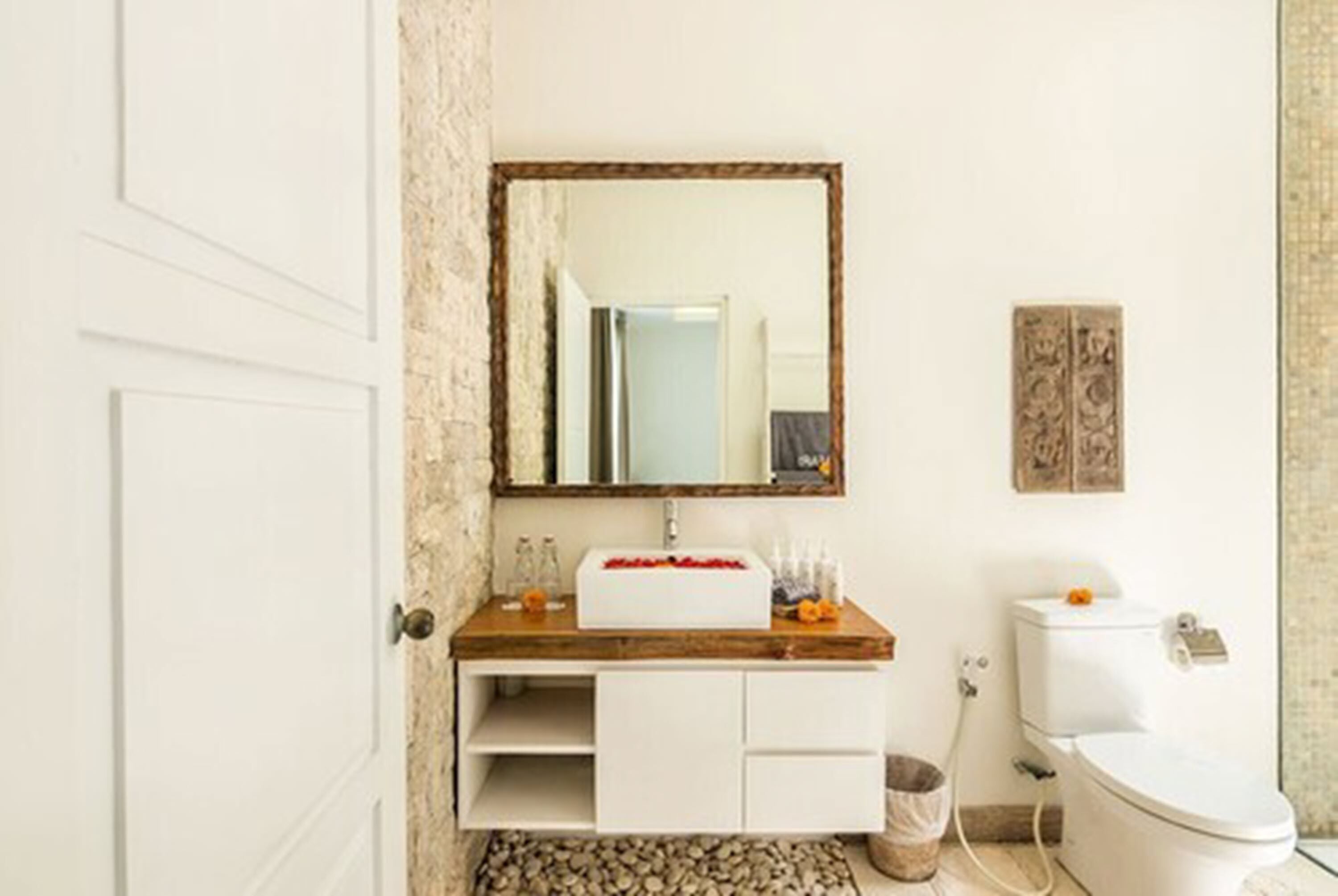 A bathroom with a wooden vanity, a white sink, a mirror, a toilet, and a small trash bin.