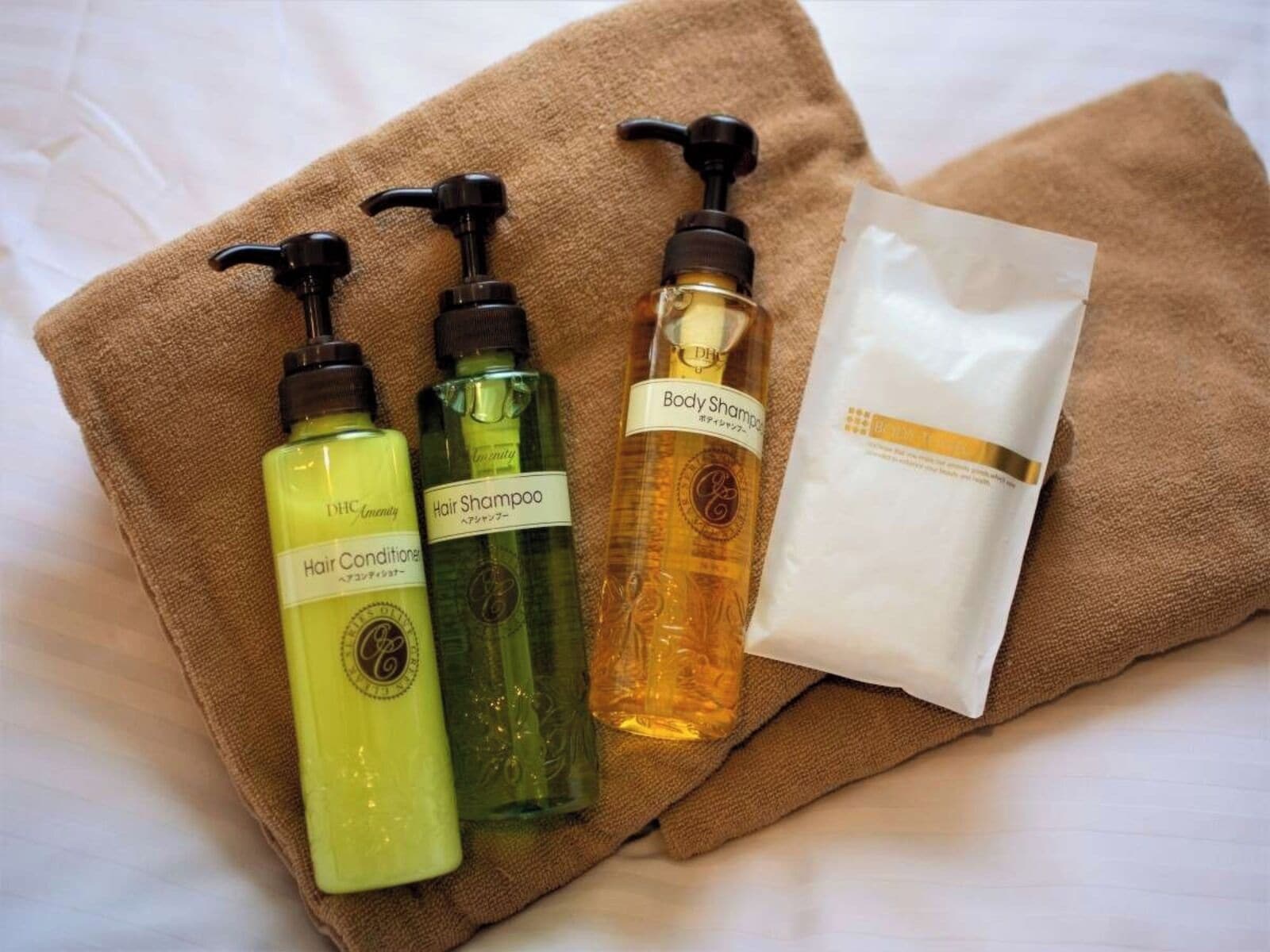 A set of toiletries on a brown towel, including hair conditioner, hair shampoo, body shampoo, and a white packet.