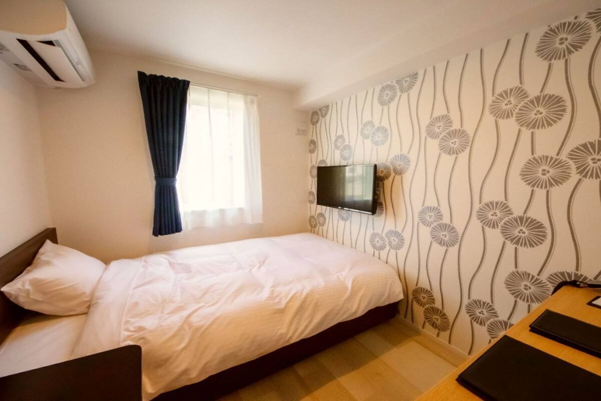 A bedroom with a bed, a TV mounted on the wall, a window with curtains, and a patterned wallpaper.