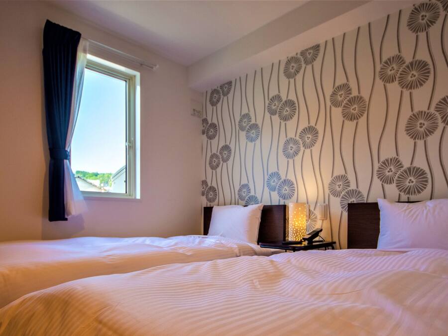 A hotel room with two beds, a window with curtains, a wall with a floral pattern, and a nightstand with a lamp.
