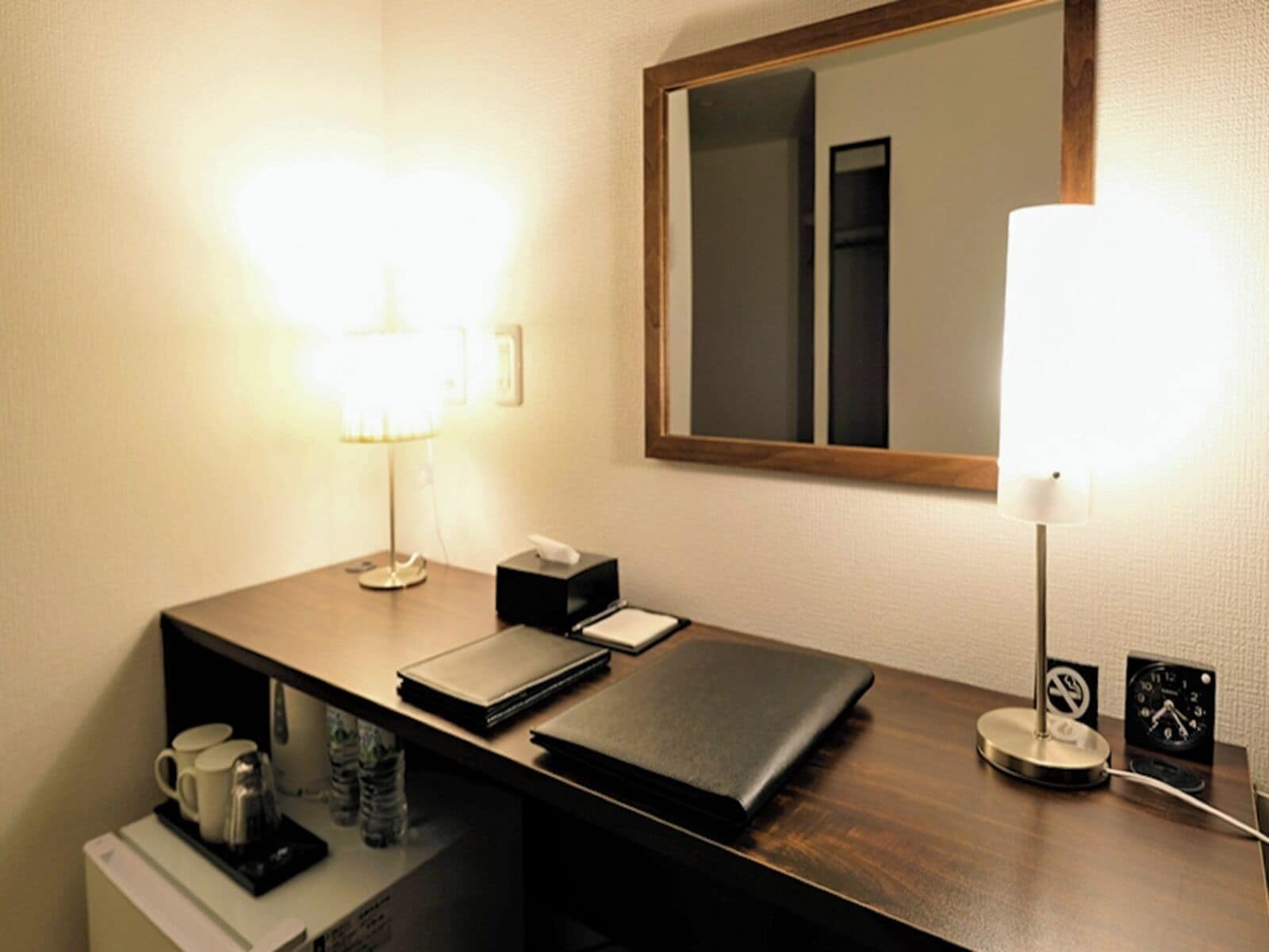 A hotel room with a wooden desk, a mirror, a lamp, a clock, and a coffee maker.
