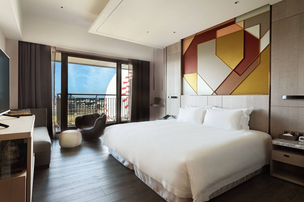 A modern hotel room with a large bed, a desk, a chair, and a balcony with a view.
