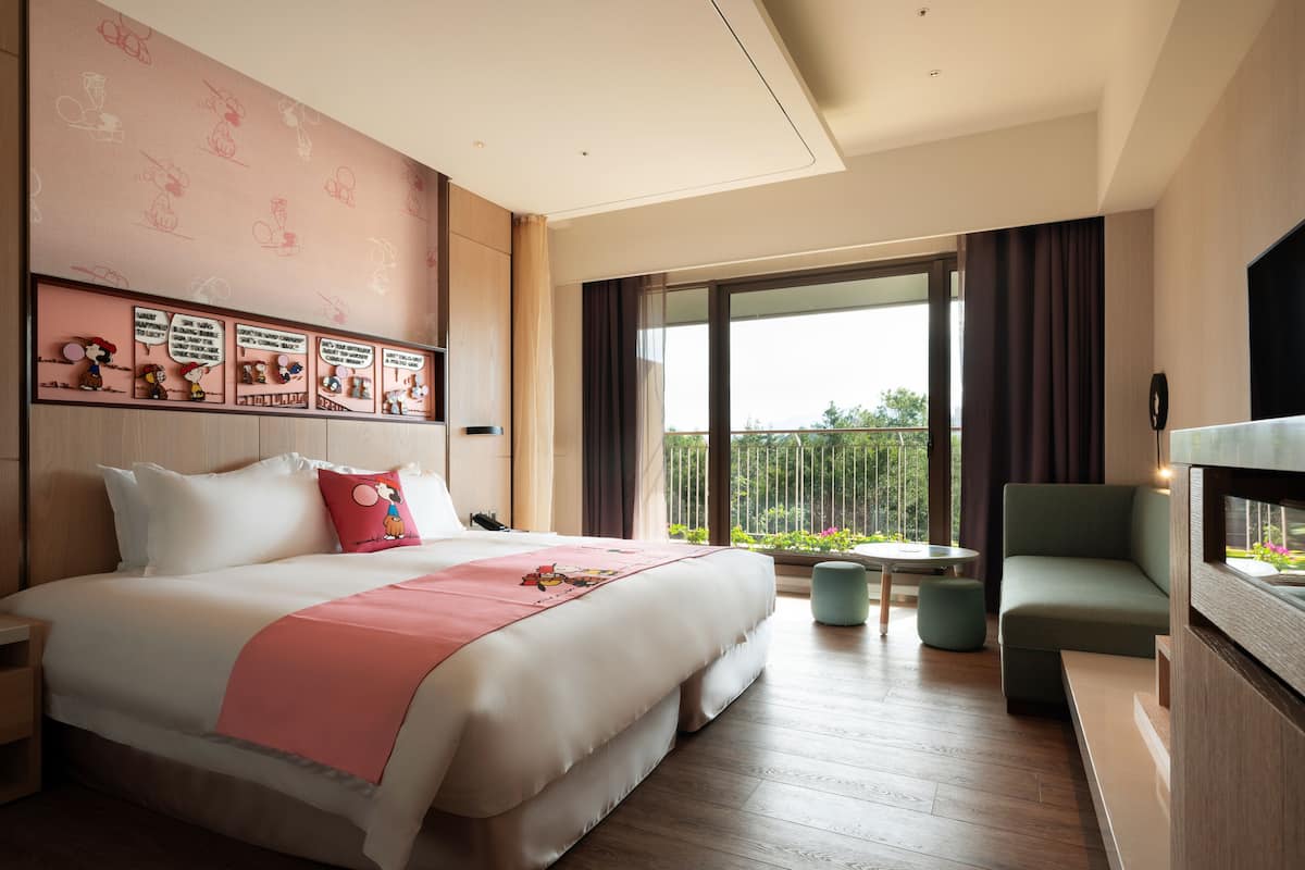 A modern hotel room with a large bed, a TV, a balcony with a view, and a small seating area.