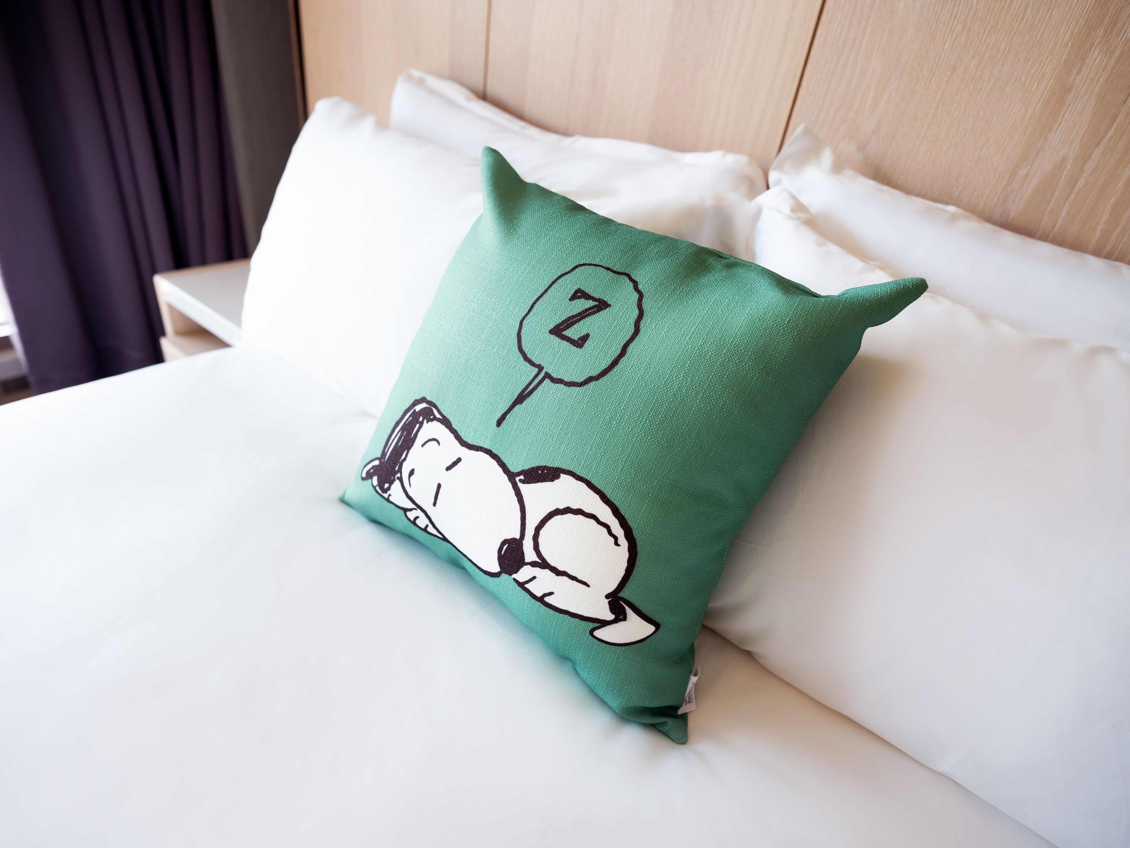 A bed with white bedding and a green pillow featuring Snoopy sleeping.