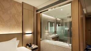 A modern hotel room with a large bed, a bedside telephone, a small bedside table, a bathroom with a glass shower enclosure, and a large mirror. - Fame Hall Garden Hotel (Taoyuan City)
