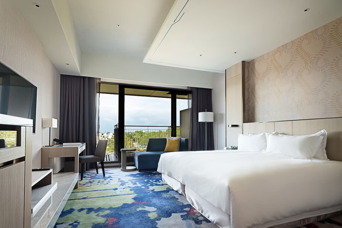 A hotel room with a large bed, a desk, a chair, a TV, and a balcony with a view.