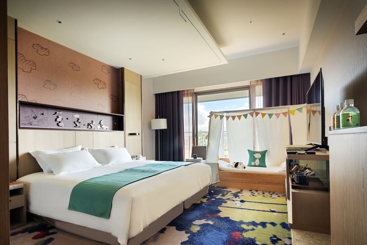 A hotel room with a large bed, a desk with a computer, a small seating area with a canopy, and a window with curtains.