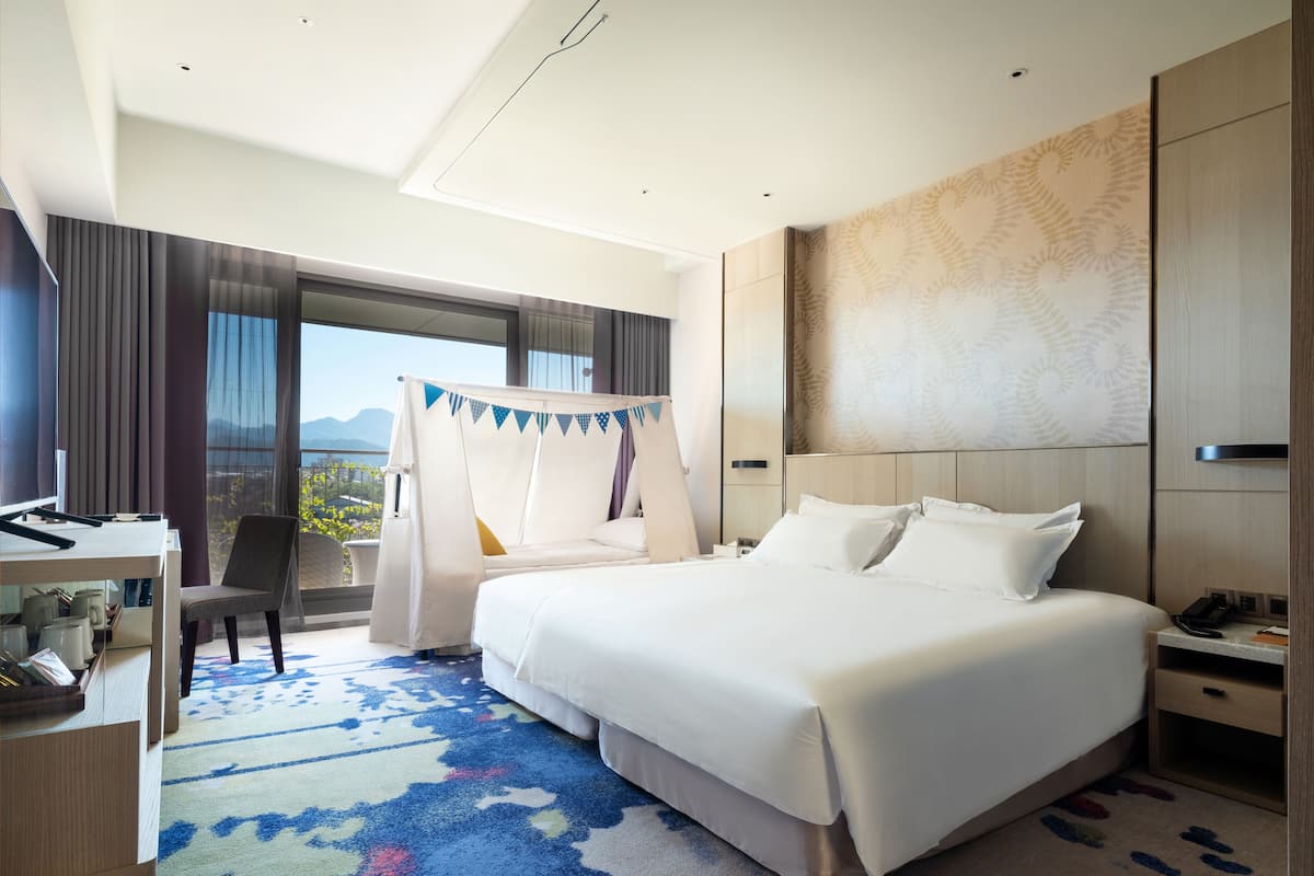 A hotel room with a large bed, a desk, and a chair. There is a view of mountains through the window.