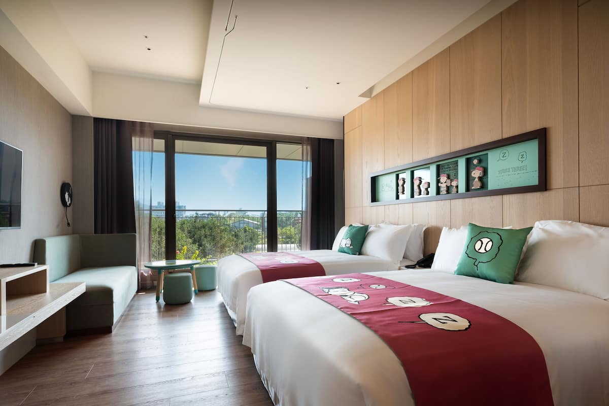 A modern hotel room with two beds, a seating area, a TV, and a balcony with a city view.